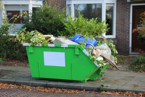 A large bright green plastic skip filled with various types of refuse, including green leafy branches, white cardboard boxes, and plastic packaging, situated on a paved area in front of residential buildings with large windows, brick walls, and surrounding greenery. The skip is positioned directly on a concrete curb adjacent to a sidewalk, with some scattered leaves and debris around it, indicative of a rubbish removal or clearance activity by Office Clearance South Kensington. The background shows garden bushes and trees, and the overall scene appears to be outdoors in a suburban or urban neighborhood in South Kensington. The skip's contents are overflowing, suggesting an active clearance service involving waste collection from domestic or small commercial premises.