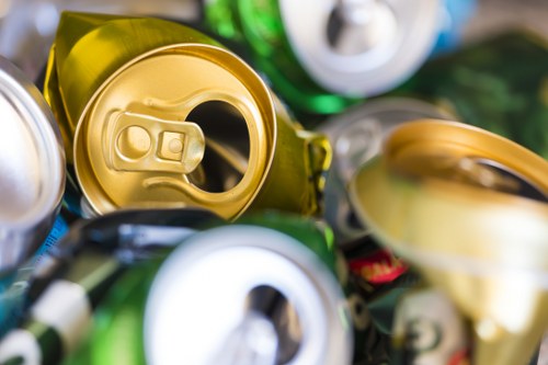 A close-up image displaying several crumpled aluminum and tin cans, primarily in silver and gold tones, piled together with some green and blue cans visible in the background. The cans are metallic, with some opened and with visible pull-tab lids, and are positioned on a surface that suggests outdoor or industrial surroundings. The cans vary in size and are stacked haphazardly, indicating waste or rubbish intended for disposal or recycling, typical of rubbish removal services offered by Office Clearance South Kensington in London’s SW7 postcode area. The environment appears well-lit with natural light highlighting the reflective surfaces of the cans, emphasizing their metallic textures. This image visually illustrates the type of waste collected during an office clearance or rubbish removal process in South Kensington, supporting the services provided by the company in clearing metal waste efficiently and responsibly.