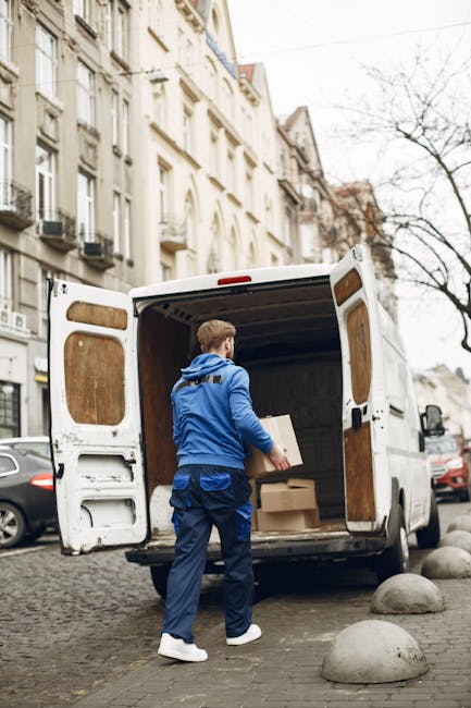 A white commercial van parked parallel to the curb along a street in front of a red brick building with decorative stonework around the windows. The van has a high roof and sliding side door, with vis