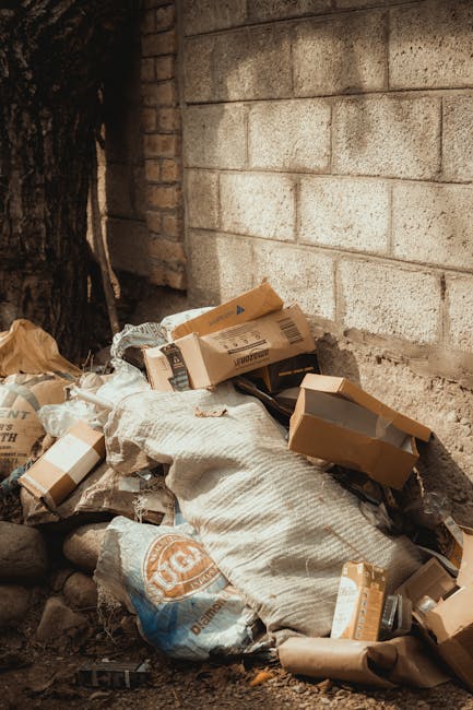 A pair of large black plastic rubbish bags and a cardboard box are placed on a sidewalk beside a black metal fence. The bags appear to be filled with waste material, and the cardboard box, which is pa