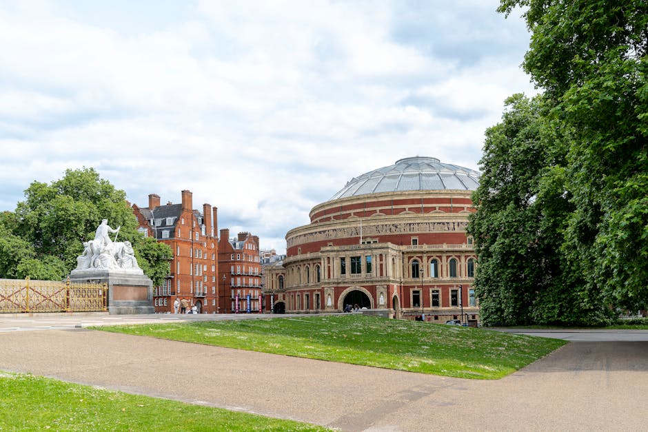 A wide view of a park area featuring a large ornate monument with a gilded spire and detailed sculptures at its base, positioned on a stone platform with decorative fencing surrounding it. To the righ