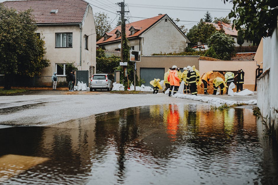 The image shows a flooded urban area between two buildings, with muddy water covering the pavement and partially filling the lower spaces of the structures. On the left, there is a white building with